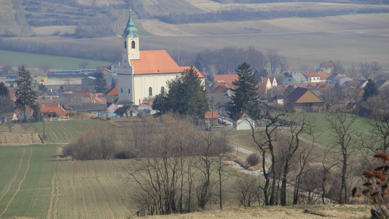 Landschaft mit Kirche und Dorf im Hintergrund, umgeben von Feldern und Bäumen.