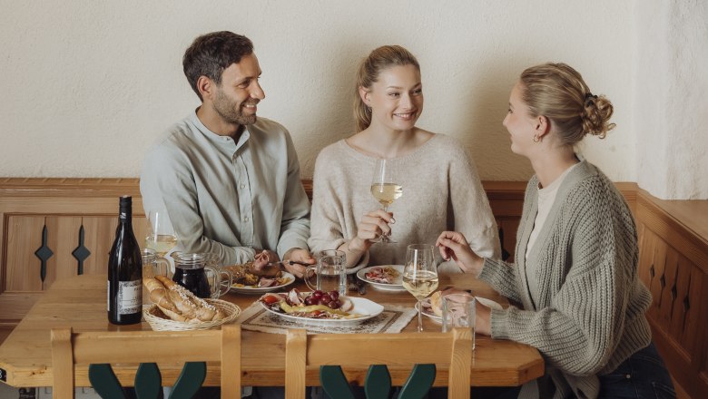 Drei Personen sitzen an einem Holztisch in einem Weinkeller und genießen Wein und Essen.