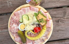 Ein Teller mit Aufschnitt, Käse, Ei, Gurke, Paprika und Brotaufstrich auf einem Holztisch. Daneben ein Glas Roséwein mit Strohhalm.