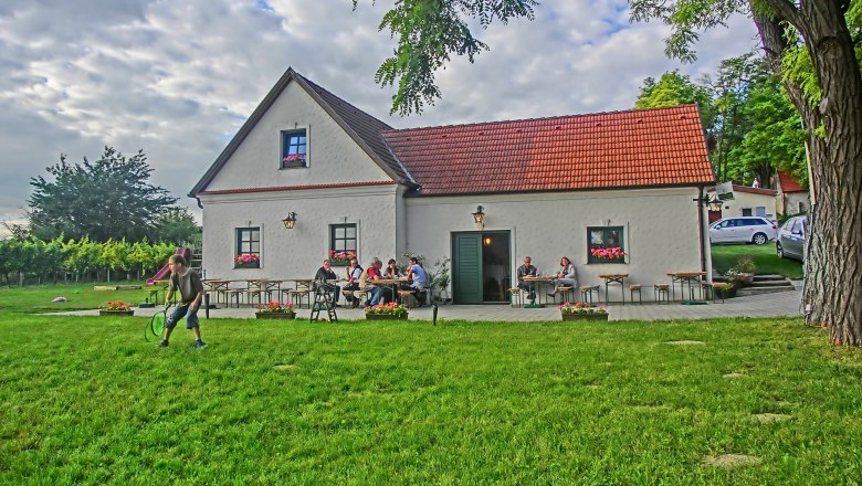 Ein traditionelles Gebäude mit rotem Dach, umgeben von grüner Wiese und Bäumen, Menschen sitzen an Tischen im Freien.