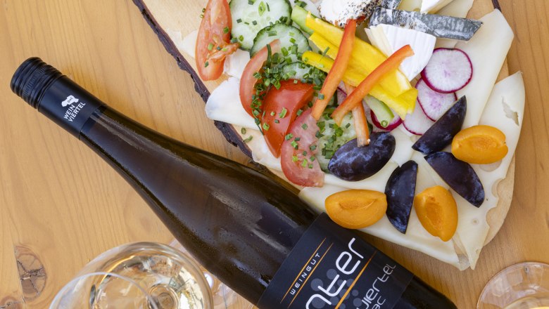 Eine Heurigenplatte mit verschiedenen K&auml;sesorten, Aufstrichen, Gem&uuml;se und einer Flasche Wein auf einem Holztisch.