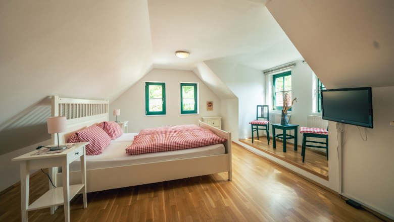 Helles Schlafzimmer mit Dachschräge, Doppelbett, Holzboden, zwei Fenstern, Tisch mit Stühlen und Fernseher an der Wand.