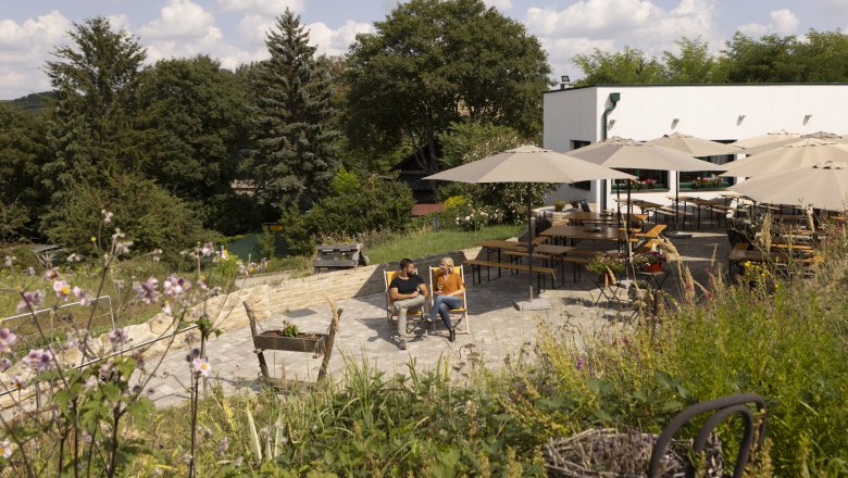 Ein Paar sitzt auf Liegestühlen in einem Garten mit Tischen und Sonnenschirmen, umgeben von Bäumen und Blumen.