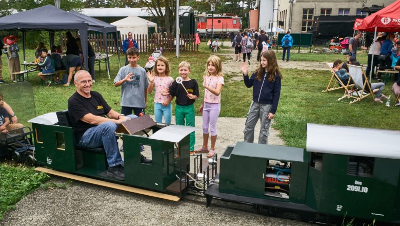 Ein Mann f&auml;hrt eine Miniatur-Eisenbahn, w&auml;hrend Kinder daneben stehen und winken. Im Hintergrund sind Zelte und weitere Besucher zu sehen.