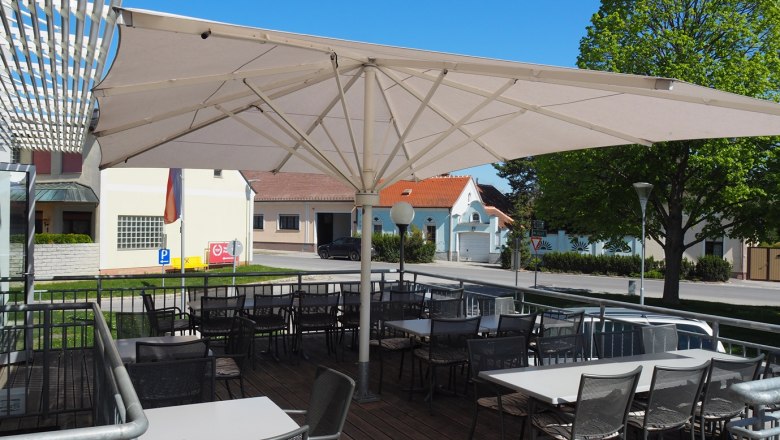 Terrasse eines Restaurants mit Sonnenschirm und leeren Tischen und St&uuml;hlen, im Hintergrund eine Stra&szlig;e und Geb&auml;ude.