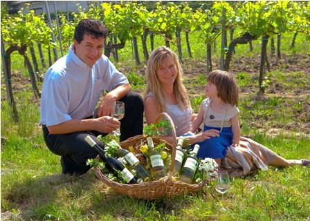 Eine Familie sitzt in einem Weinberg neben einem Korb mit Weinflaschen.