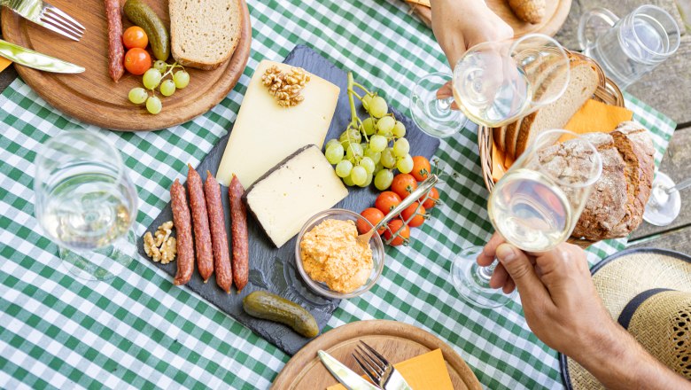 Picknicktisch mit Käse, Brot, Wurst, Trauben und Wein auf kariertem Tischtuch.