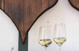 Zwei Weingl&auml;ser mit Wei&szlig;wein auf einem Holztisch vor einer herzf&ouml;rmigen Holzdekoration.