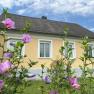 Ein gelbes Haus mit weißen Fensterrahmen, umgeben von blühenden rosa Blumen im Vordergrund.