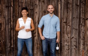 Zwei Personen stehen vor einer Holzwand, eine Frau mit Weinglas und ein Mann mit Weinflasche.