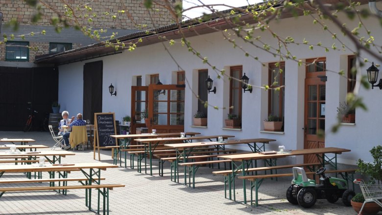 Innenhof einer Buschenschank mit leeren Holzb&auml;nken und einem Tisch, zwei Personen sitzen an einem Tisch, ein Spielzeugtraktor steht daneben.