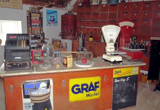 Innenansicht eines historischen Dorfladens mit Waage, Registrierkasse und alten Werbeschildern.