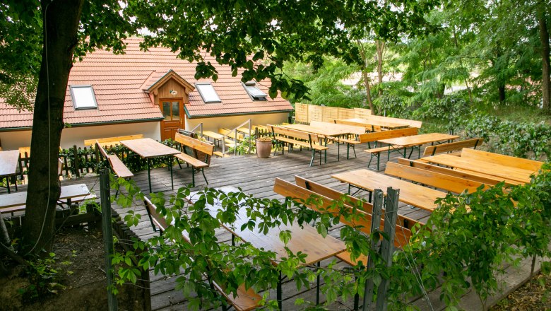 Ein leerer Biergarten mit Holzb&auml;nken und Tischen unter B&auml;umen, neben einem Geb&auml;ude mit rotem Ziegeldach.