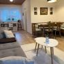 Gemütliches Wohnzimmer mit grauem Sofa, Esstisch und Stühlen, Bücherregal und Fenster mit Gardinen.